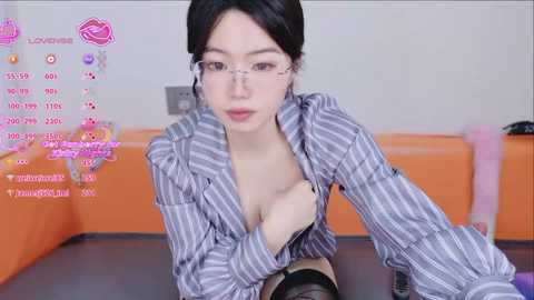 A video of an Asian woman with pale skin and dark hair in glasses, wearing a partially unbuttoned striped shirt, sitting on a bed with orange sheets. The background includes a pink heart icon and social media metrics.