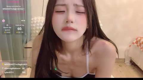 Video of a young Asian woman with long black hair, wearing a black camisole, eyes closed, and flushed cheeks, in a dimly lit bedroom.