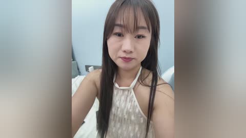 Video of a young Asian woman with long black hair and fair skin, wearing a white sleeveless top, sitting indoors with a blurred background.