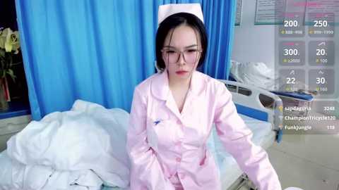 Video of an East Asian woman in a pink nurse's uniform and cap, standing in a hospital room with blue curtains, white bed, and medical charts.