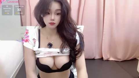 Video of an East Asian woman with long black hair, wearing a white crop top exposing a black bra, sitting on a bed with pink curtains in the background.