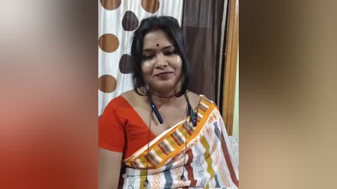 Video of a middle-aged Indian woman with medium skin tone, wearing a traditional orange saree with a striped border. She has a neutral expression and a small bindi on her forehead. Background includes polka-dotted curtains and a brown door.