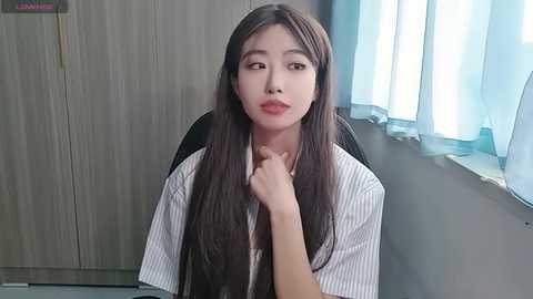 A video of an East Asian woman with long black hair, wearing a white pinstriped shirt, sitting in an office chair by a window with light blue curtains.