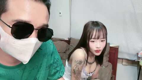 A video shows an Asian woman with long black hair and tattoos, wearing a white lace bra, and a man in a green shirt and mask.