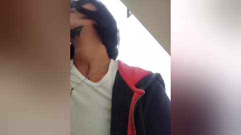 Video of a young woman with medium brown skin, long black hair, wearing a white ribbed shirt and a black and red zip-up jacket, captured from a low angle in a dimly lit hallway.