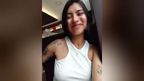 A video of a young woman with medium-length black hair, light skin, and a smile. She wears a white sleeveless tank top, showcasing tattoos on her arms and shoulders. The background shows a modern, dimly lit room with a wooden shelf.