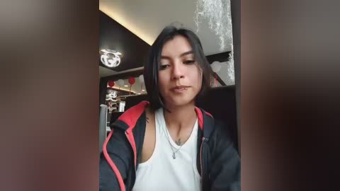 A video of a young woman with medium-length dark hair, wearing a black jacket over a white tank top, standing in a dimly lit room with exposed brick walls and modern decor.