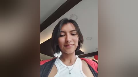 Video of a young woman with short, dark hair, wearing a white tank top and a red jacket, smiling in a modern indoor setting.