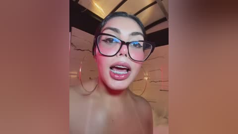 Video of a young woman with medium brown skin, wearing black-rimmed glasses, large hoop earrings, and a black top. She has dark hair pulled back, and her lips are slightly parted, revealing braces. The background is dimly lit with wooden beams.