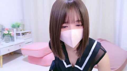 Video of an Asian woman with shoulder-length straight brown hair, wearing a black sailor-style top with white trim and a pink face mask. She is seated in a pastel-pink room with a white desk, potted plants, and a pink beanbag chair in the background.
