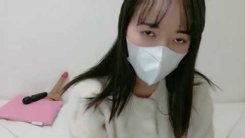 Video of an Asian woman with long black hair, wearing a white mask and pink nail polish, holding a pink book, in a plain white room.
