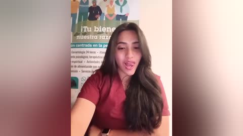 Video of a young woman with long black hair, fair skin, wearing a red t-shirt, making a tongue-out expression in front of a colorful poster in Spanish.