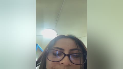 Video of a young woman with light brown skin, brown hair, and glasses, taken from a low angle, showing her upper body and face. Background features a white ceiling and a round light fixture.