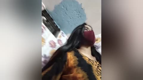 A video shows a woman with long black hair, wearing a maroon mask and a tie-dye top, standing in a room with a blue ceiling and floral wallpaper.