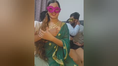 Video: A woman with long wavy hair in a green saree with gold embroidery, wearing a pink masquerade mask, sitting on a sofa. Behind her, a man in a white shirt and a woman in a grey sweater cuddle.