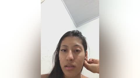 A video of a young woman with medium brown skin, long black hair, and dark eyes, wearing a neutral expression, adjusting her hair with one hand in a white-walled room with a ceiling light.