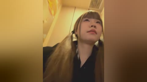 Video of a young woman with long, straight, light brown hair in pigtails, wearing a black shirt, standing in a dimly lit hallway with beige walls. She has a serene expression and is gazing upward.