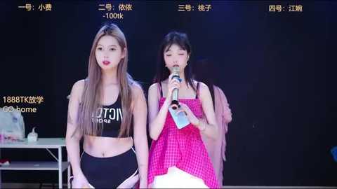 Video of two young Asian women, one in a black crop top and high-waisted shorts, the other in a pink dress, singing into microphones on stage.