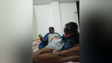 Video of two people: a young man in a blue shirt, and a woman with long black hair and a blue patterned shirt, lying on a bed in a dimly lit room.