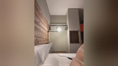 A video of a modern bedroom with a wooden headboard, white bedding, a sleek black wall-mounted lamp, and a minimalist gray wardrobe in the background. The scene is dimly lit.
