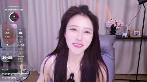 Video of a smiling East Asian woman with long black hair, wearing a pink top and pearl necklace, in a cozy living room with a floral arrangement and a clock.