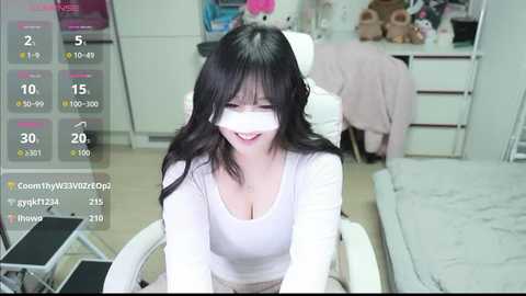 A video of a smiling Asian woman with long black hair, wearing a white mask, white top, and beige pants, sitting in a white chair in a cluttered room with toys and furniture.