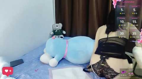 A video of a light-skinned woman lying on a bed with a plush toy and a black bra visible. The room has a blue sheet and dark curtains.