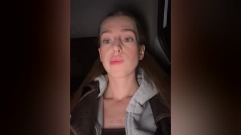 Video of a Caucasian woman with light skin, straight brown hair, and fair complexion, wearing a denim jacket over a light-colored shirt, sitting in a dimly lit car.