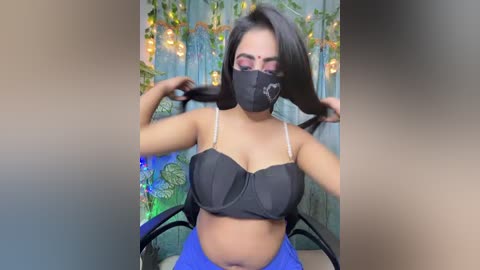 Video of a young woman with long black hair, wearing a black face mask, black bra, and blue pants, sitting in a chair against a colorful, festive backdrop.