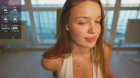 Video of a young woman with fair skin, long brown hair, wearing a white tank top, looking thoughtful. Background shows a blurred indoor setting with large windows and an outdoor view.