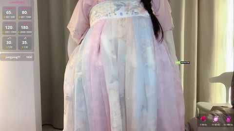 Video of a person from behind, wearing a pastel-colored, flowing dress with a high waist, standing in a beige-walled room with a window and a chair.