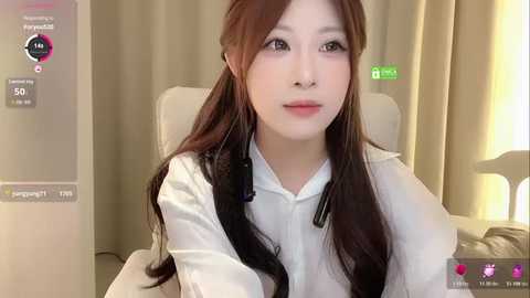Video of an East Asian woman with long, straight brown hair, wearing a white blouse, sitting in a beige chair, with a green light on a wall in the background.