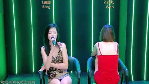 Video of two women on stage, one in leopard print dress, the other in red dress, facing audience, singing into microphones.