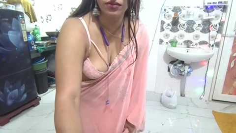 Video of a South Asian woman with medium skin tone, wearing a pink saree revealing a lacy bra, in a cluttered, tiled bathroom with a sink and colorful decor.