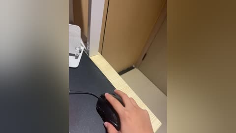 A video of a person's hand holding a black computer mouse on a beige wooden desk. The background features a beige wall and a white electrical outlet with a cord plugged in.