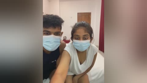 Video of a young South Asian couple wearing masks, sitting on a bed with white sheets, in a simple, dimly lit room with white walls and a wooden door.