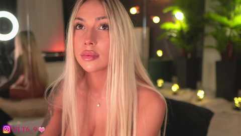 Video of a blonde woman with long hair, wearing a strapless top, in a dimly lit room with greenery and glowing lights in the background.