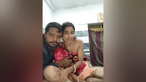 A video of an Indian couple with the man wearing a black shirt, and the woman in a red saree, sitting close in a modest, white-walled room.