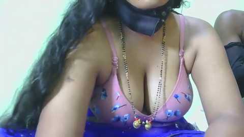 Video of a South Asian woman with long black hair, wearing a pink bra with blue designs, a black choker, and a gold necklace, against a blurred background.