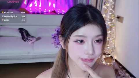 Video of an Asian woman with long black hair, wearing purple floral hairpins, smiling softly. Background features a white couch, a string of fairy lights, and a purple pillow.