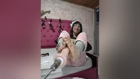 Video of a young woman with glasses, wearing a pink and white tutu, seated on a bed with pink tufted headboard. Her legs are bound by a large white and pink orthopedic boot, with a metal contraption attached to her ankle.