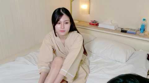 Video of a young Asian woman with long black hair, wearing a beige robe, sitting on a white bed in a minimalist hotel room.