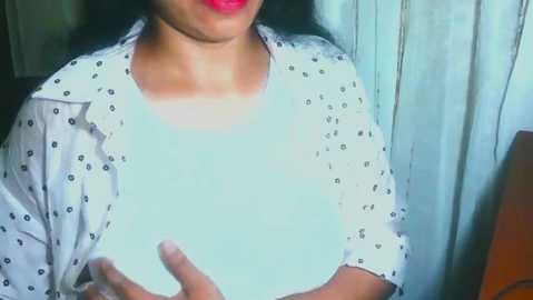 Video of a woman with medium skin tone, wearing a white blouse with black polka dots, holding a white shirt against her chest. Background features a light-colored wall with vertical stripes and a wooden chair to the right.