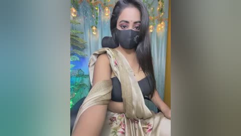 Video of a South Asian woman with medium brown skin, long black hair, and a black face mask, wearing a black bra and a floral saree. She poses against a colorful, nature-themed wall mural with fairy lights in the background.