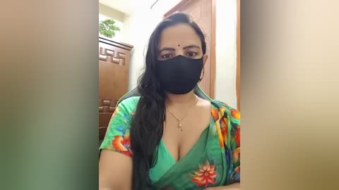 A video of a South Asian woman with long, dark hair, wearing a green saree with colorful floral patterns, black face mask, and glasses, indoors near wooden furniture and a green plant.