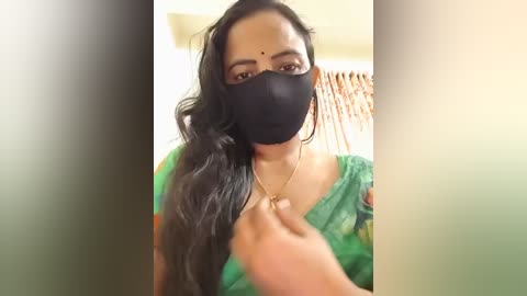 A video of a South Asian woman with long, wavy black hair, wearing a green saree, black face mask, and gold earrings. She's being touched affectionately on the cheek by a hand. The background is blurred, showing a wooden wall.