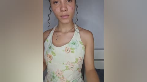 Video of a young woman with curly hair, medium skin tone, wearing a floral-patterned tank top, sitting against a plain, light-colored wall.