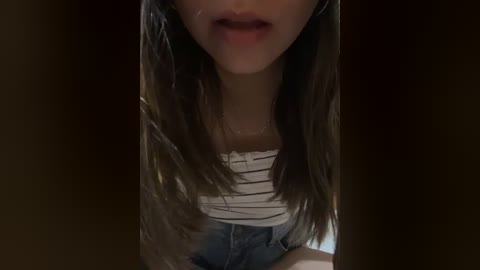 Video of a young woman with long, straight brown hair, wearing a white striped top and blue jeans, partially obscured by a dark background.