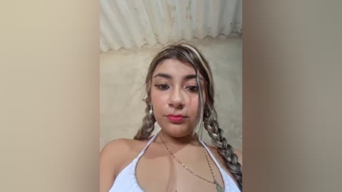 Video of a young woman with light skin, long braided hair, and a white bikini top, standing against a textured beige wall.