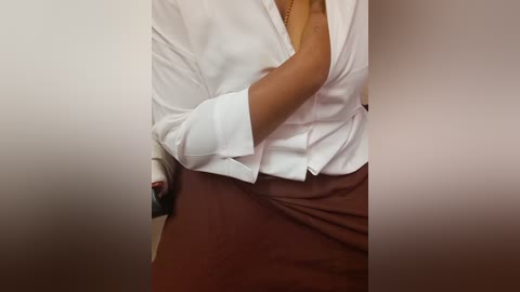 A video of a woman's torso, wearing a white blouse unbuttoned to reveal a small cleavage, and a brown skirt. She holds a smartphone in her left hand. Background is a plain, beige wall.